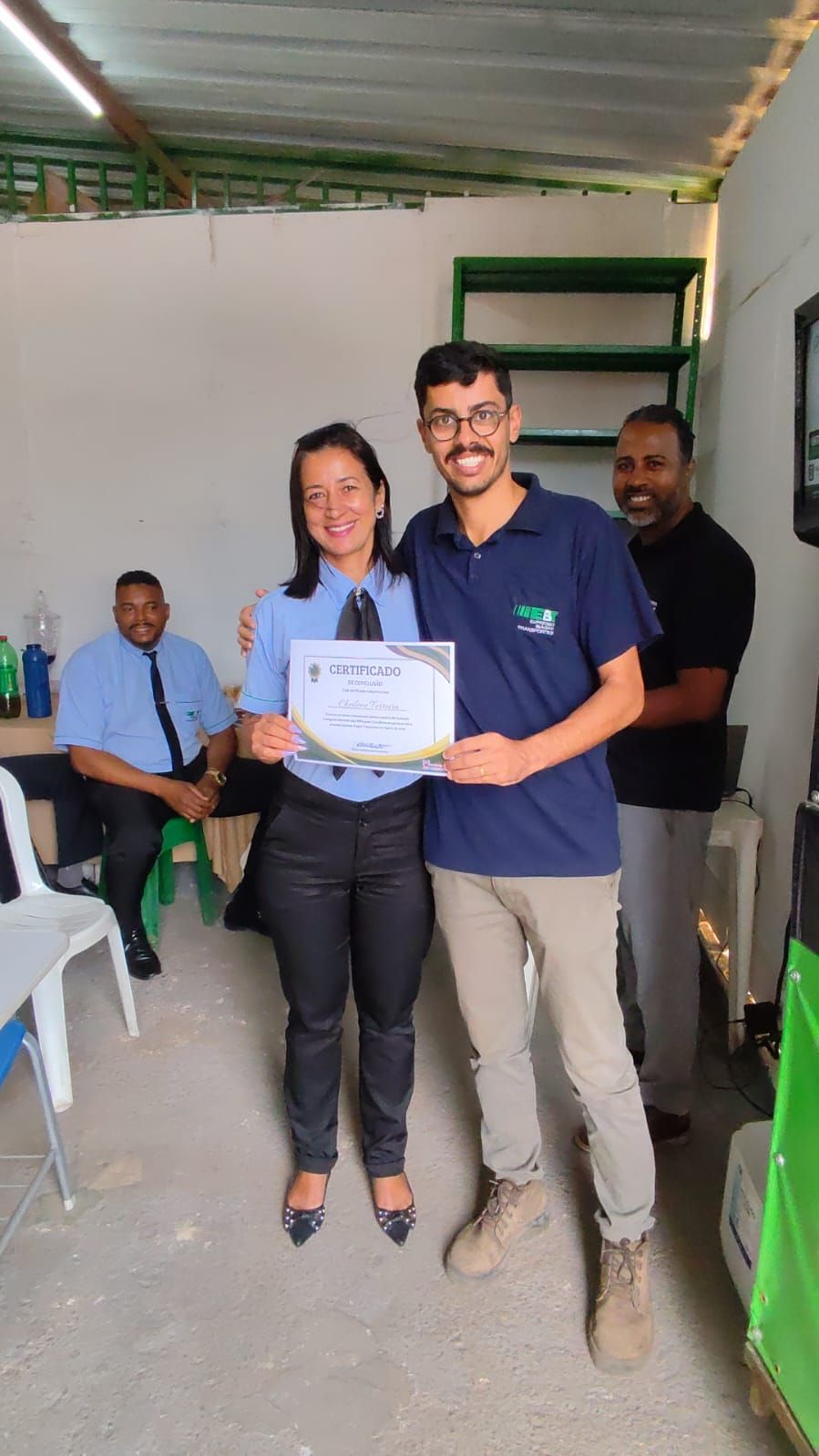 Entrega de certificado de capacitação em uma oficina, com colegas ao fundo